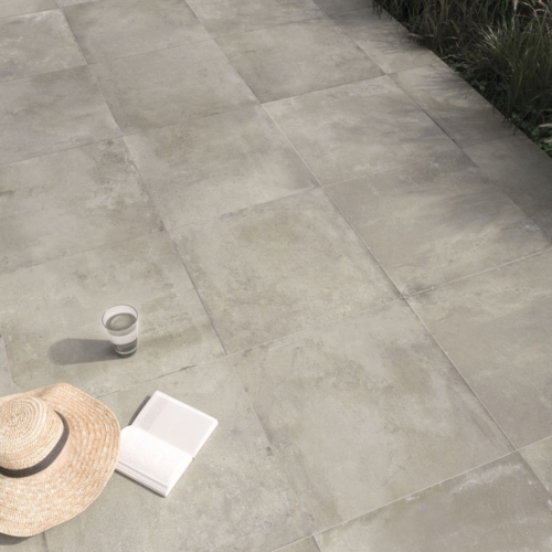 Strohhut, aufgeschlagenes Buch und Glas Wasser auf gefliester Terrasse neben Gras.