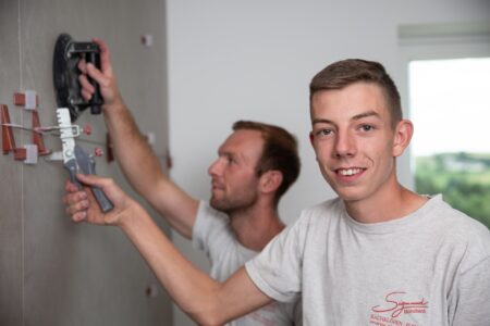Zwei Männer bei der Wandarbeit, einer hält ein Werkzeug, beide in grauen T-Shirts.